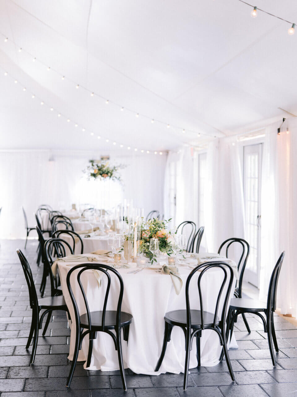 Tented reception with string lights, black bentwood chairs, and white linens—strong detail shot